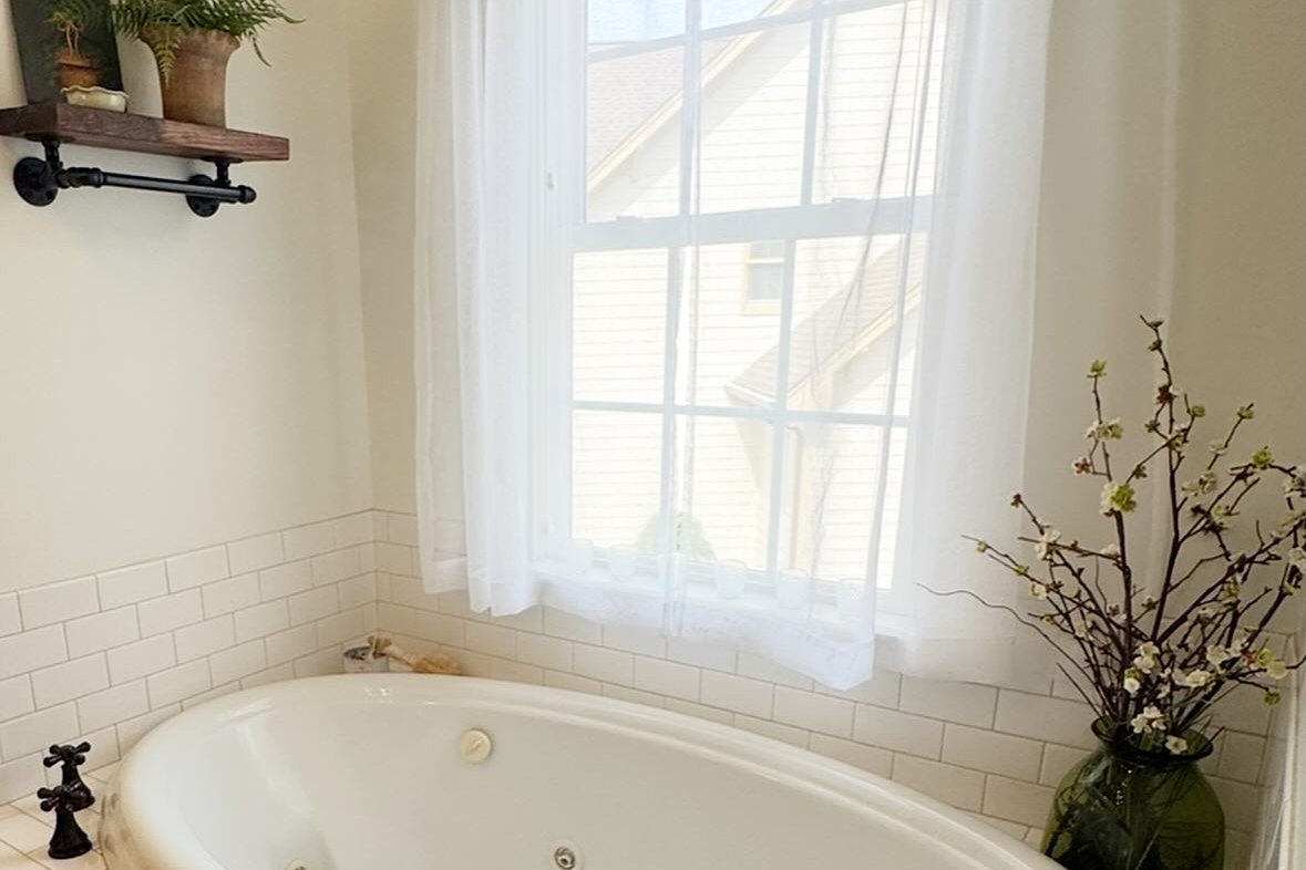 A soaking tub beside a sunny window with sheer curtains and a folded towel